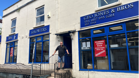 A man in shorts and a dark polo shirt stands at the entrance to a white building, with blue shop frontage advertising 'TAPS', 'SHOWERS' and 'GEO JONES & BROS' above three consecutive shop windows. There is a red 'Closing down sale' sign in the main window to the right.