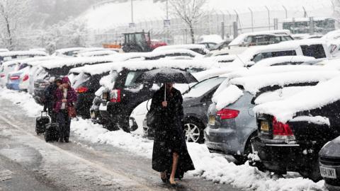 Woman walking through a car park in heavy snow carrying an umbrella