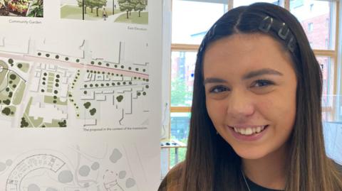 Emily has long hair and is smiling while standing next to some of her design plans on a banner in an exhibition room