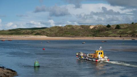 A boat on a river with sand dunes and grass in the background.