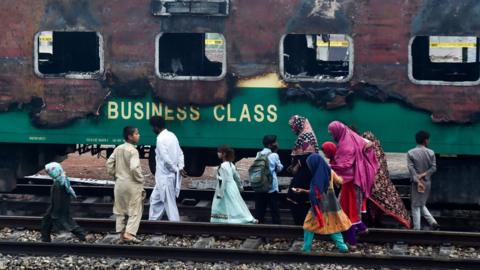 A family walks passed the charred remains of a train carriage