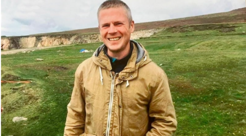 Jim O'Neill, a man with grey hair, wearing a brown jacket, standing on grass, near a cliff.