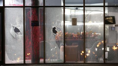 Large window panels have been smashed. The office on the other side of the glass panels is covered in sprayed red paint