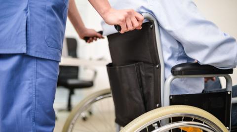 A person in blue medical scrubs pushes a wheelchair with a man wearing a light blue shirt sitting in it. 