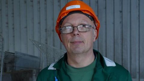 Andy Simms inside a silver coloured metal factory at a sugar beet factory. He is wearing a bright orange hard hat, glasses and a dark green jacket over a brighter green sweatshirt. 