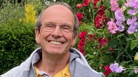 Stephen, who has short grey hair, back from a receding hairline, is wearing glasses and is smiling broadly. He has a grey sweatshirt over a yellow top and is standing in front of a hedge with red, pink and mauve sweet peas growing on his left.