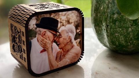 A wooden cube with round edges sits on a white side table. On the face that points forward is a picture of an old woman and an old man - both white - smiling and laughing. 