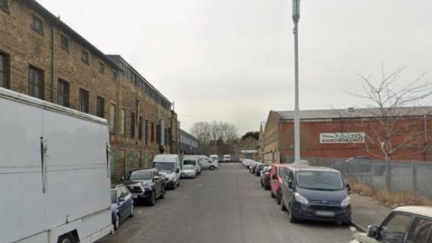 A street in Bradford which has an old mill building on one side and a red brick commercial property on the other. Cars line each side of the street. 