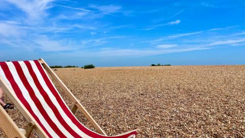 View of a shingle beach with a red and white deckchair under a blue sky