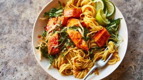 A bowl filled with noodles, tofu, spinach, a wedge of lime and a fork.