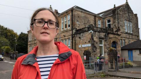 Karen Reid stands in front of Milngavie Primary School. She is wearing a stripy top, and red jacket and glasses with a neutral expression. The primary school is an old sandstone building, slightly dirty with age.