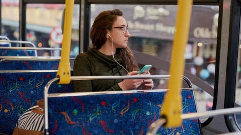 A young woman wearing glasses looks out the window of a bus while listening to her phone using headphones.
