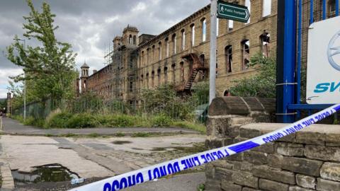 Blue and white police tape with the shell of a burnt-out stone three-storey mill building in the background.