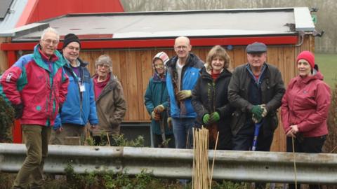A group of middle-aged men and women are standing next to each other in outdoor jackets holding shovels and sticks. They are all smiling at the camera.