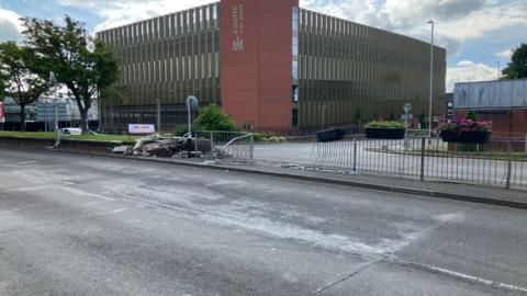 A street scene shows a damaged brick wall and police tape, suggesting a recent crash. A tree and the "Castle Car Park" can be seen in the background.
