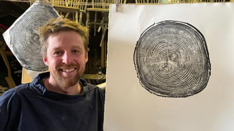 A man in a navy blue jumper with a beard standing beside a parge print showing the rings of a fallen tree