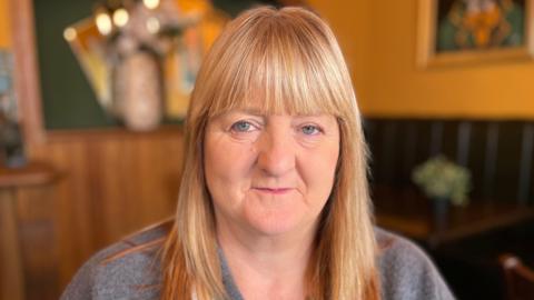 Joan has long blond hair and is wearing a jumper