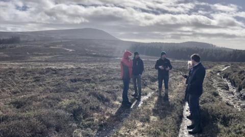 Four people standing in a group on a trail on a flat expanse of mossy wetland, with a hill and a forest in the background.