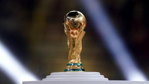 The FIFA World Cup Trophy. it shows two sculpted human figures stretching upward, holding the Earth aloft. The design meant to symbolise the global reach of football and the unity of nations. it sits on a white pedestal
