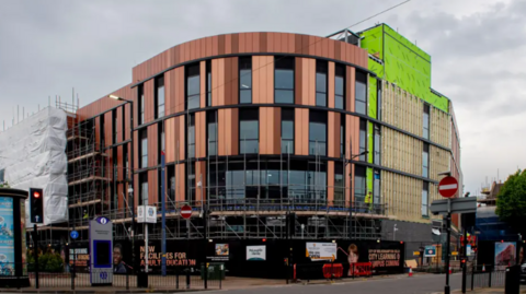 The large orange and black curved building stands on a corner. It has a green and gold section on the right hand side of the image. There are some advertising hoardings and small scaffolding at ground level.