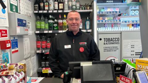 Corner shop manager stood behind a shop counter.