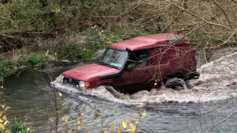 A red 4x4 drives along a section of a Hampshire chalk stream open to traffic
