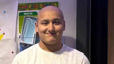 Ethan is standing with a toothless grin for the camera. He has on a white t-shirt and has close cropped hair. It is a head shot of him.