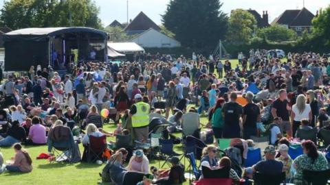 Crowd on grass area in a park at a festival with a large stage to the left.