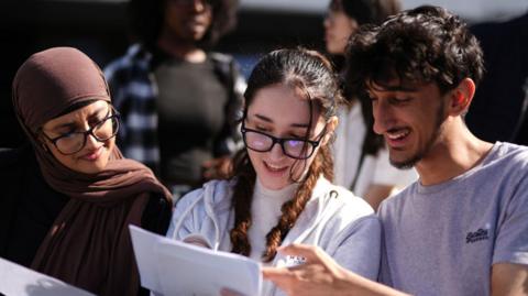Two female students and a male student look down at an exam results sheet they've just opened. 