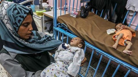 Woman holding an apparently malnourished child as another one lies in a bed
