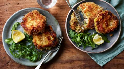 Two grey plates on a wooden table filled with golden crumbed fishcakes and green leaves