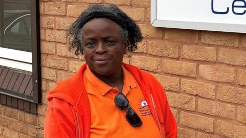 Naomi has medium length black and grey hair and is wearing a hairband, orange t-shirt, red jacket. She is stood in front of a brick wall and looking at the camera.