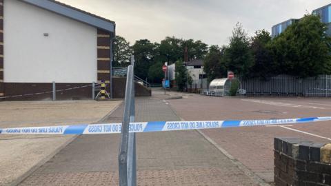 A car park next to a warehouse building, cordoned off with blue and white police tape. It's a cloudy day.