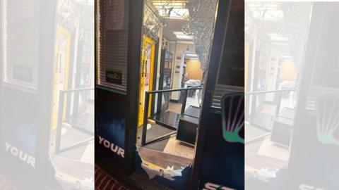 A smashed front door of a shop. Inside, there are yellow doors and what appears to be a black cabinet on the floor.