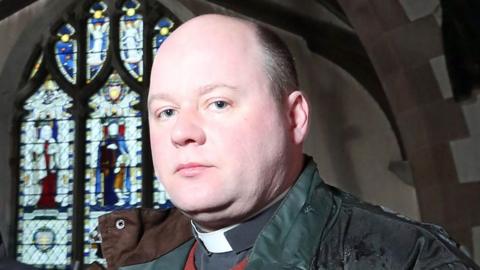 Adrian Robert McLaughlin standing in front of a stained-glass window wearing a dog collar, jumper and a waterproof coat.