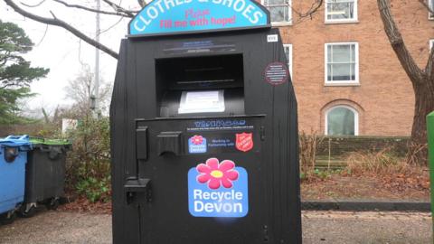 A black bin that says "clothing & shoes". Behind is to the left are two bins, one blue and one black, to the right is a tree and a brown brick building.