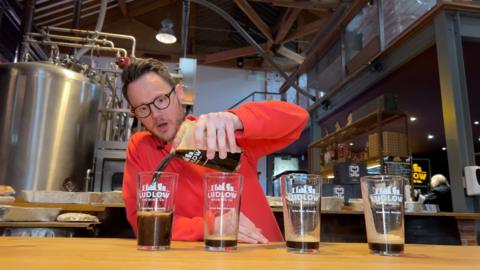 a man with a beard and glasses in a red-ish jumper pouring beer into a pint pot at a brewery