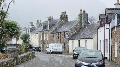 Village scene showing road and parked cars with snow falling