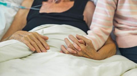 An older patient holds a young relatives hand in bed 