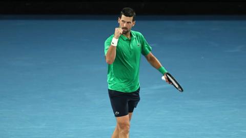 Novak Djokovic celebrates on the court. He is wearing a green shirt and black shorts. 