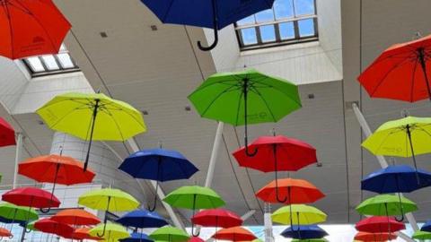 A number of colourful open umbrellas hanging from the roof of a shopping, seen from below. They are in bold colours: red, blue, yellow and green with black handles. 