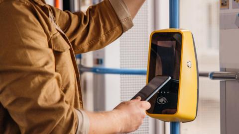 A close up of a person wearing a beige jacket tapping their phone on a contactless machine attached to a pole on a bus.