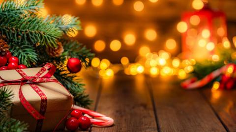 A wrapped present is sat under the branches of a Christmas tree and there are lights in the background