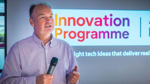 Digital Jersey chief executive Tony Moretta holds a microphone while he speaks at an event for the Innovation Programme launch. A sign with the project's name on it is behind him.