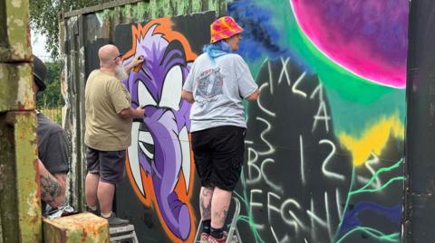 Two artists painting brightly-coloured pictures. One painting is of a purple elephant with tusks. The other painting has letters and numbers