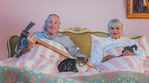 Keith and Marj are sat up in a bed with pink and white striped bedding. There is a cat sat between them. Both of them are holding building tools. The wall behind them is pink and has a hole in it exposing wooden panels. On the same wall is a gold mirror.