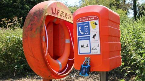 Orange life ring secured in an orange case on a pole. Next to it is a box that reads "rescue line" and has a padlock on it