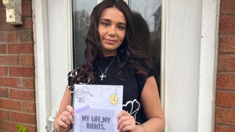 Photograph of Shannon, a young woman with long dark hair, stood outside her front door. She's wearing a black T-shirt with white flowers on it, and has a silver cross necklace on. Shannon is looking into the camera and smiling slightly. She's holding a white laminated booklet, which has 'My Life, My Rights' written on it in black lettering. 
