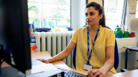 A teacher wearing a yellow shirt and a blue "staff" lanyard sits at her classroom desk in front of a computer. You can't see what is on the computer screen. There is a large window behind her and plants and books on the windowsill. 