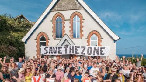 Hundreds of people waving their arms in the air in front of the Methodist chapel with a banner draped across the front of it saying "Save the Zone".